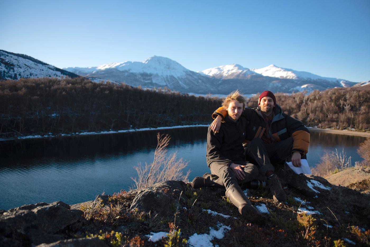 Los dos fundadores sentados sobre una roca frente a una laguna, con bosque y montañas nevadas de Aysén al fondo