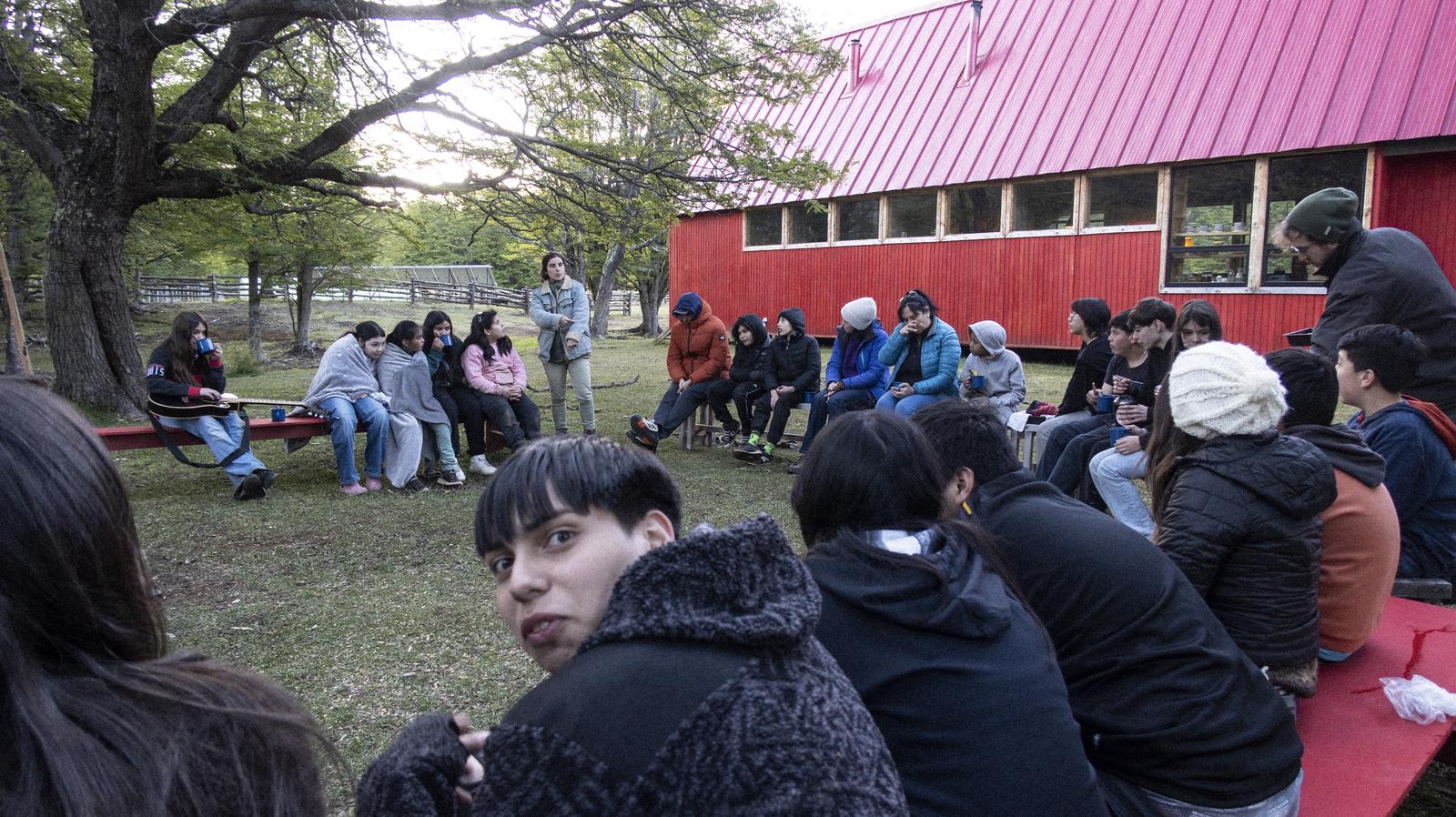 Estudiantes conversan sentados en el pasto frente al Refugio Escuela rojo