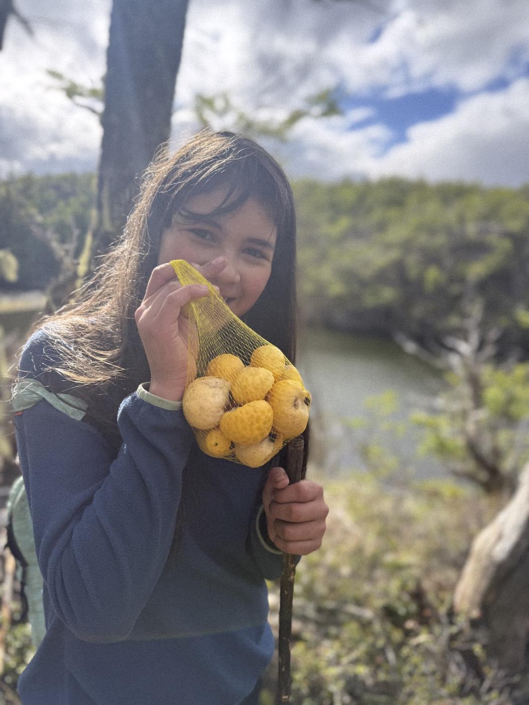 Una estudiante sonríe mostrando una malla con frutos recolectados, con el lago y el bosque al fondo