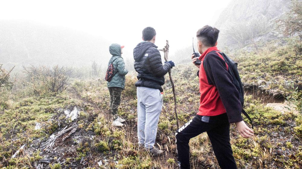 Tres estudiantes observan la ladera entre la niebla, con bastones y mochilas