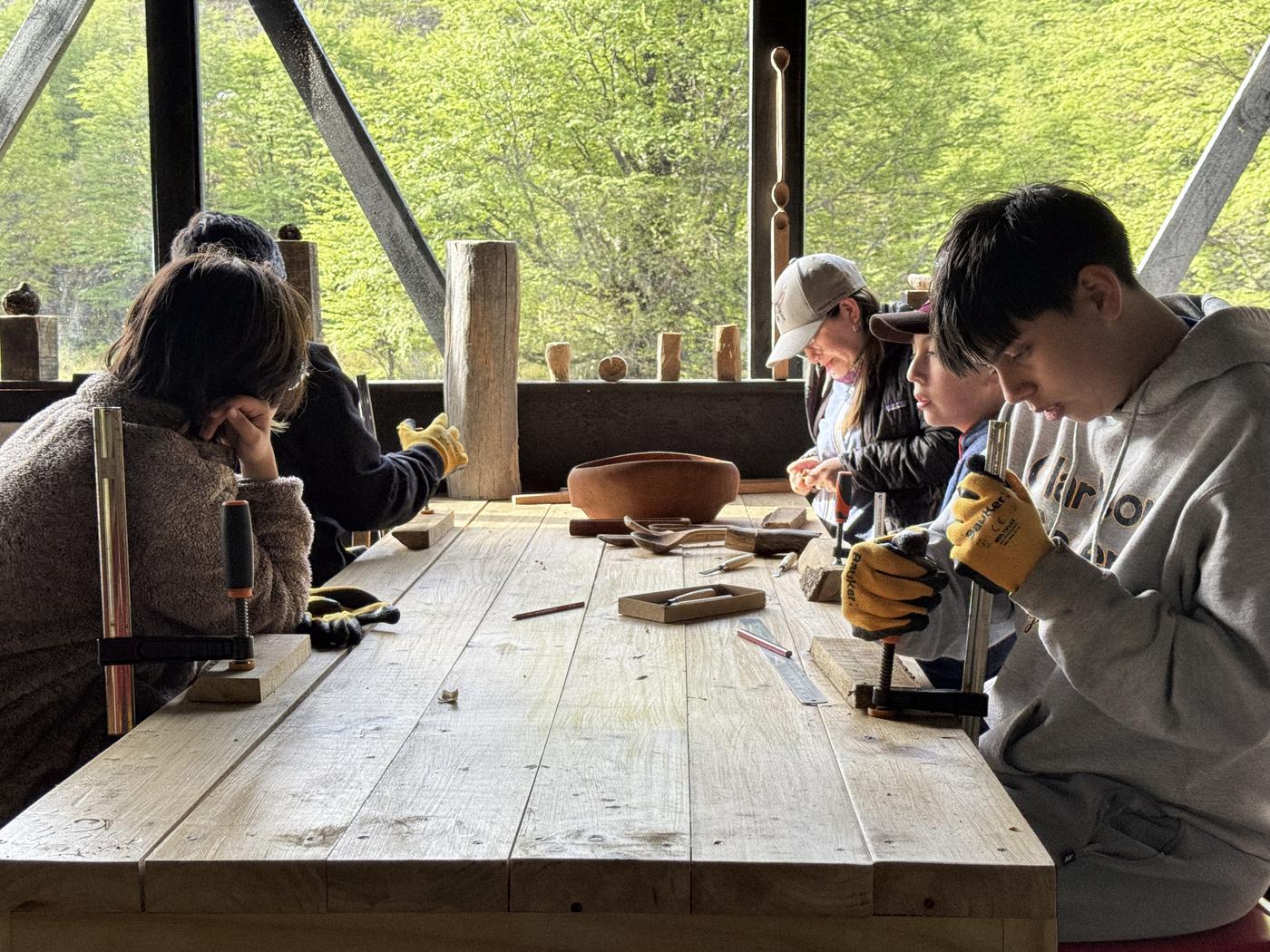 Estudiantes trabajan la madera en la mesa larga del taller, con el bosque al fondo