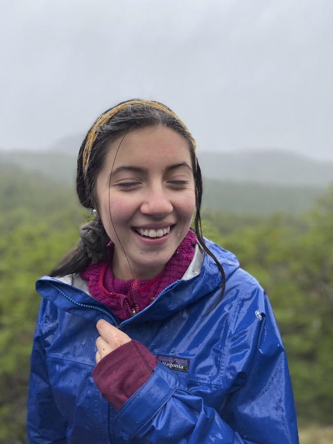 Una estudiante con chaqueta azul sonríe bajo la lluvia con el valle de fondo